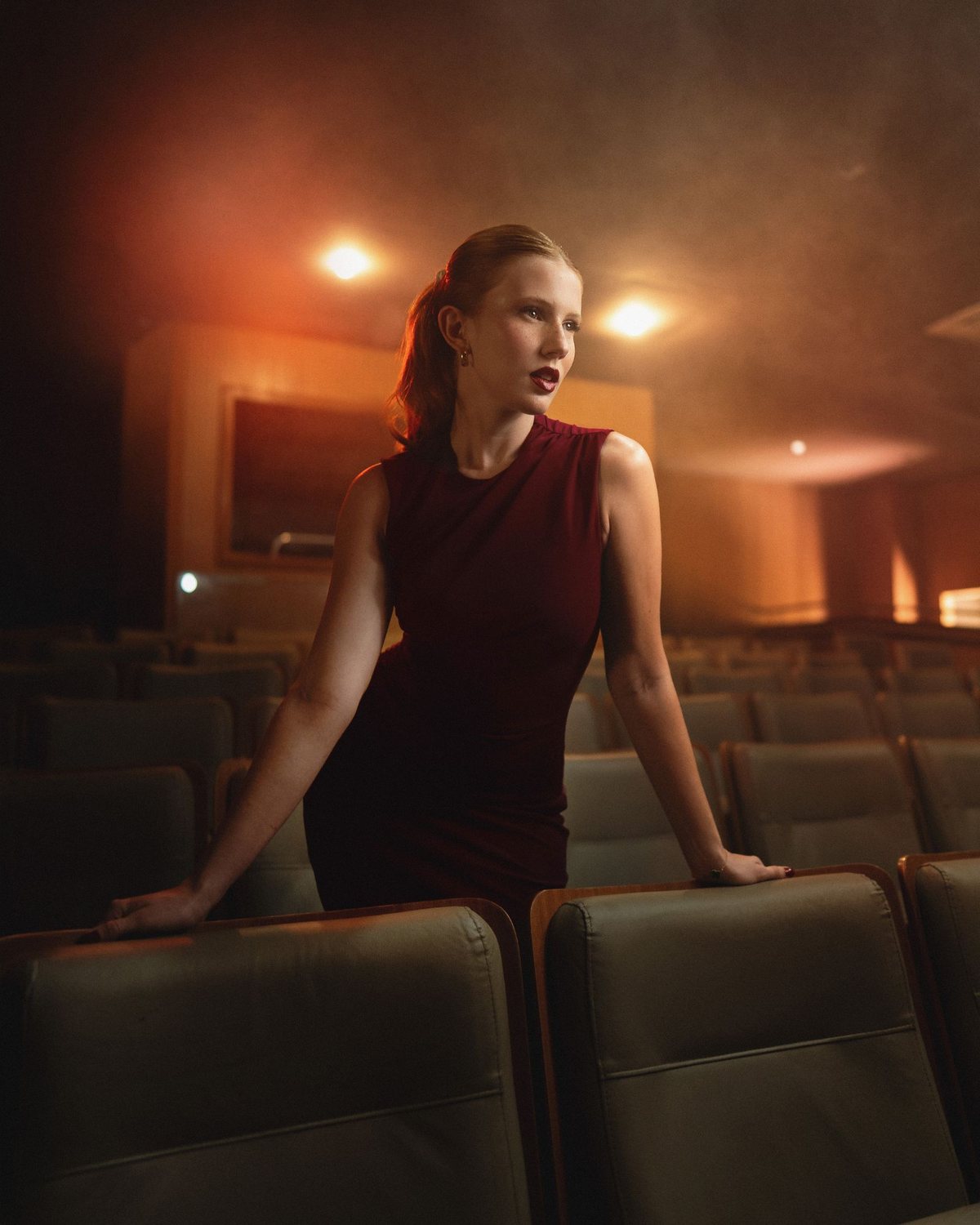 Retrato em teatro: mulher de vestido vinho entre as poltronas, com fumaça cenográfica e luz quente