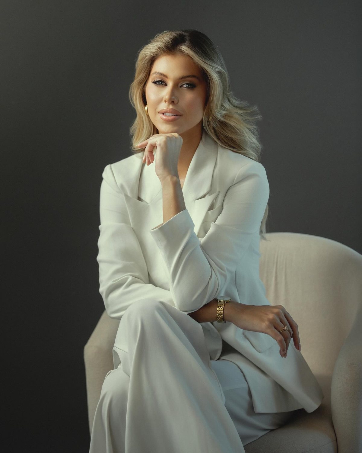 Retrato corporativo de mulher de terno branco sentada em poltrona, com a mão no queixo, em fundo cinza escuro, Vitória-ES