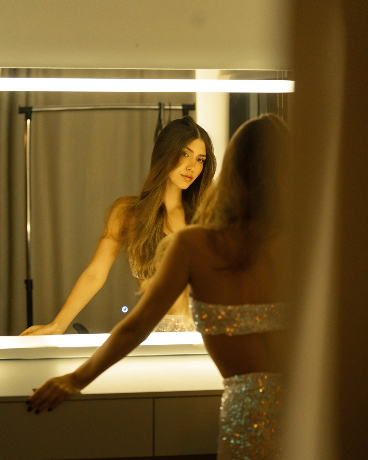Mulher diante do espelho iluminado do camarim, em tons dourados