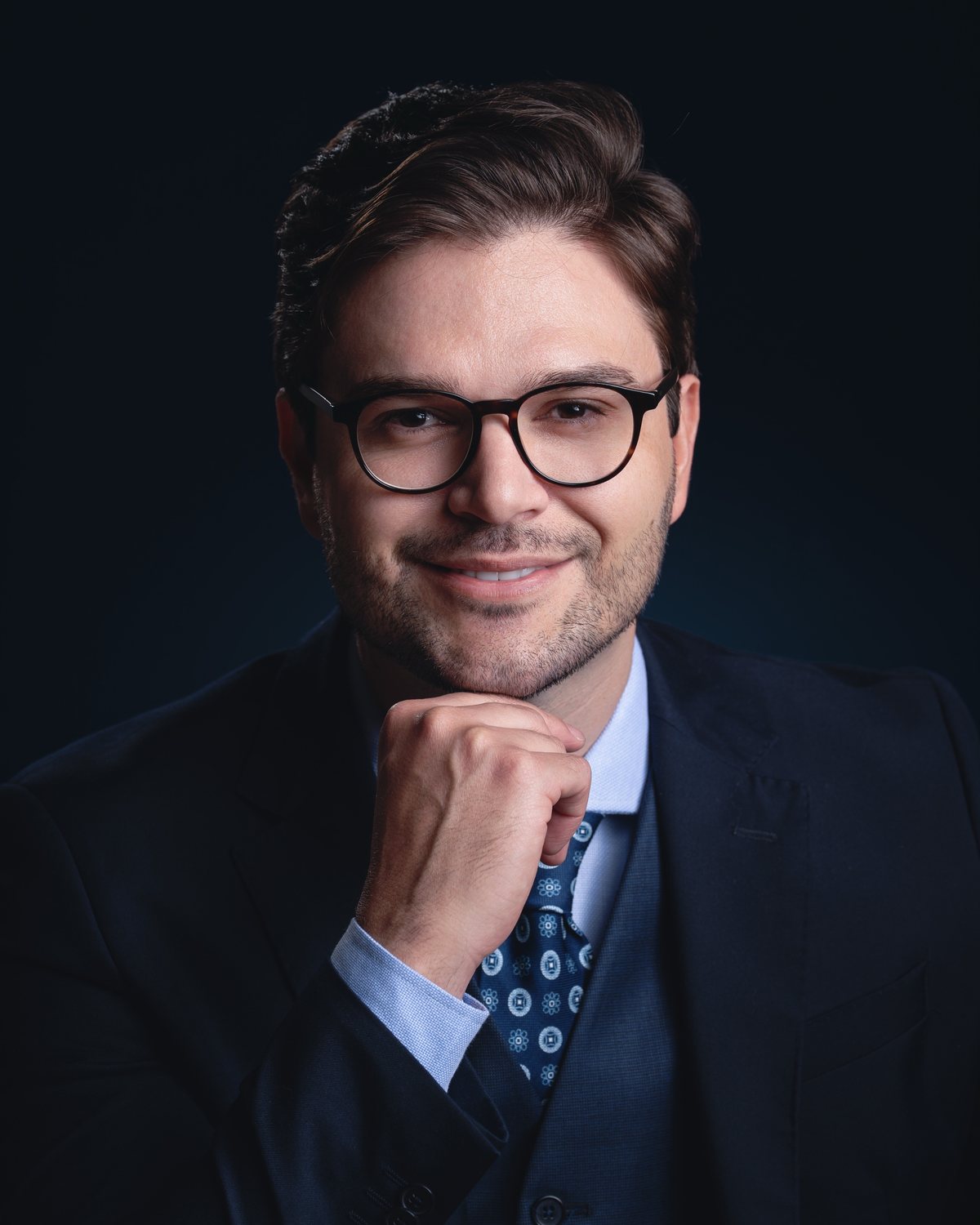 Retrato para LinkedIn: homem de óculos, terno e gravata azul, com a mão no queixo